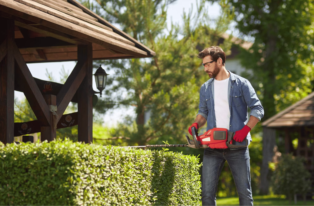Professional landscaper wearing protective safety glasses using a hedge trimmer to shape a green garden hedge